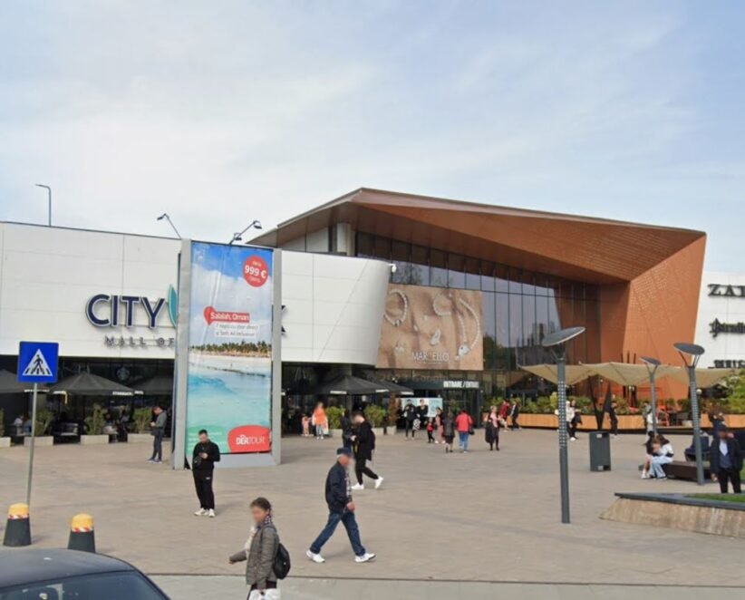 People walk and gather outside a modern shopping mall with a white façade and a copper-colored angular entrance. retailers and banners are visible outside the entrance, City Mall.