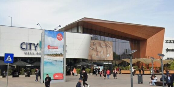 People walk and gather outside a modern shopping mall with a white façade and a copper-colored angular entrance. retailers and banners are visible outside the entrance, City Mall.