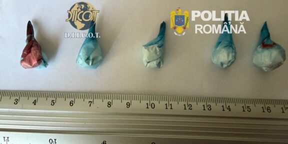 Five small wrapped parcels laid out on a ruler for scale; logos of DIICOT and Romanian Police in the background, suggesting a drug seizure display.