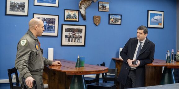 Two men talk at a wooden table in a blue-walled room with framed aviation photos and a mounted hawk between them.