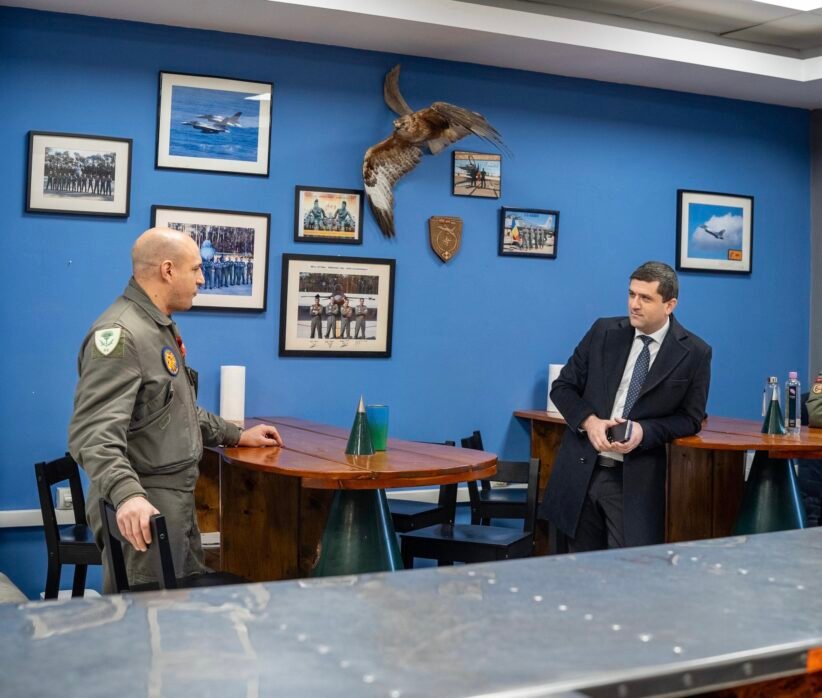Two men talk at a wooden table in a blue-walled room with framed aviation photos and a mounted hawk between them.