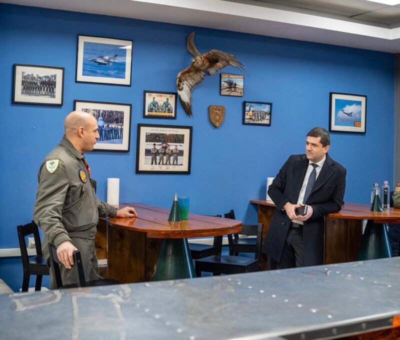 Two men talk at a wooden table in a blue-walled room with framed aviation photos and a mounted hawk between them.