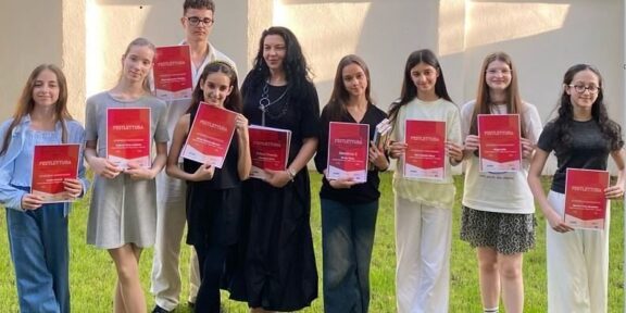 Eleven students and a teacher pose on a lawn, each holding a red certificate, celebrating a ceremony outdoors.