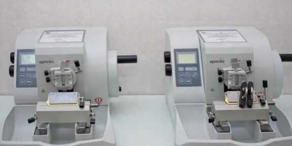Two compact gray laboratory instruments with control panels sit side by side on a lab bench, each with a sample clamp and cutting area.