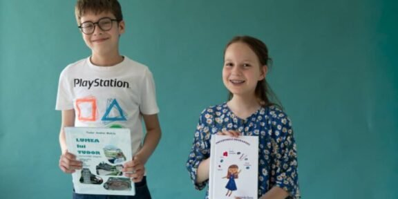 Two smiling children, a boy with glasses in a white PlayStation t-shirt and a girl in a blue floral dress, hold books in front of a teal wall.