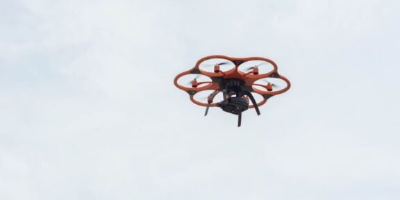 Orange hexacopter drone with six propeller arms hovering in a pale blue sky