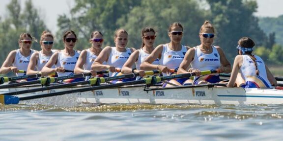 Women’s eight rowing team in a white-and-blue boat racing on a lake, each athlete with oars raised and wearing sunglasses and team uniforms.