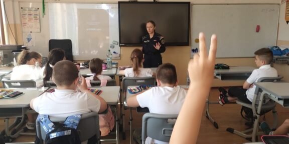 Uniformed officer giving a talk to elementary students in a classroom; a student in the foreground raises a hand to answer.