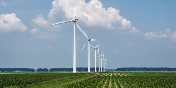 Row of tall white wind turbines stretching across a flat green farm under a blue sky with fluffy clouds
