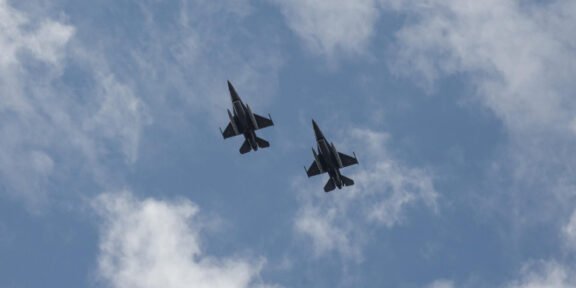 Two fighter jets in formation flying through a partly cloudy blue sky.