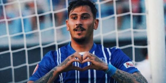 Soccer player in a blue jersey forming a heart shape with his hands in front of a goal net.
