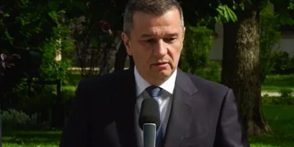 Man in a dark suit speaks into a microphone outdoors, with trees in the background.