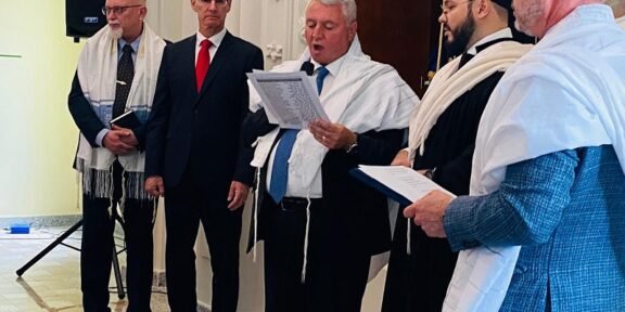 Man in a suit reads from a sheet as four other men in white prayer shawls stand nearby in a formal ceremony setting.