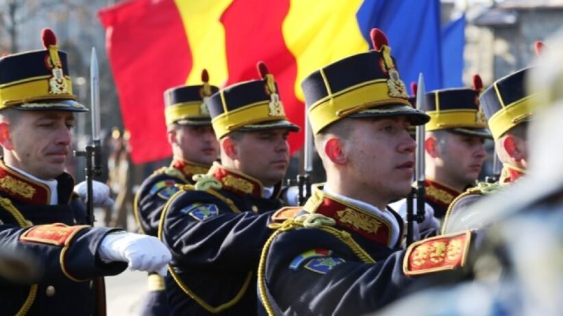 Group of uniformed soldiers marching with rifles during a parade, with red, yellow, and blue flags in the background.