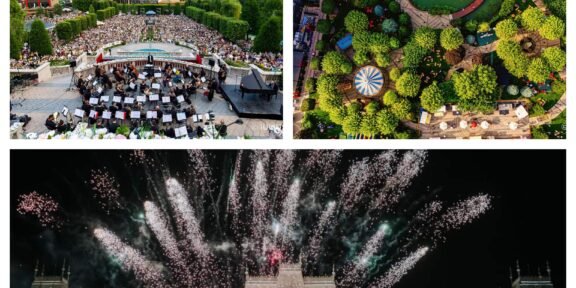 Outdoor orchestra concert in a city square with a large audience and surrounding trees.