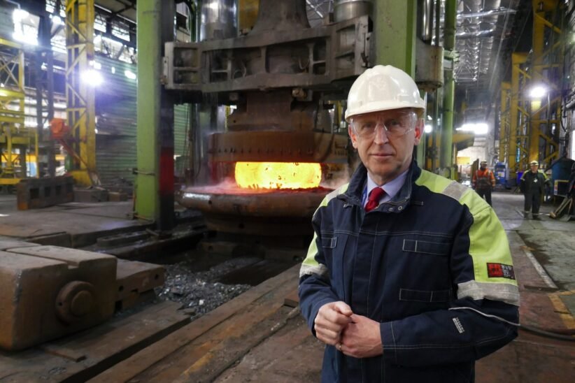 Industrial foreman in hard hat and safety jacket beside a glowing molten metal furnace in a factory.