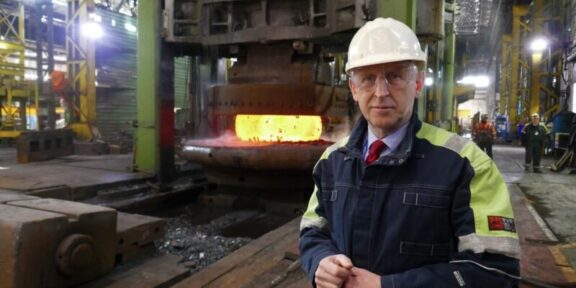 Industrial foreman in hard hat and safety jacket beside a glowing molten metal furnace in a factory.