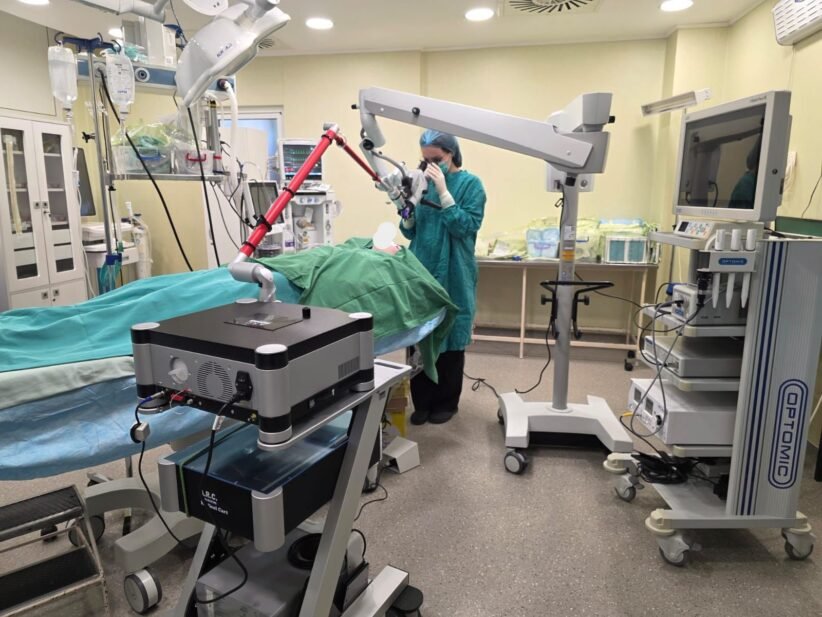 Surgeon in teal scrubs operates a robotic-assisted system over a patient on an operating table in a bright OR.