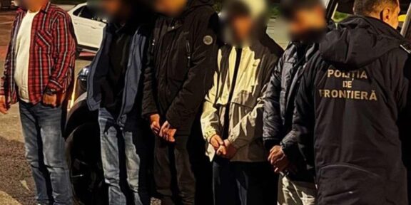 Detainees with hands bound stand in a line at night beside a border police officer wearing a jacket that reads 'Poliția de Frontieră'.