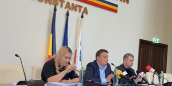 Press conference with three officials seated at a long desk, microphones in front of them, flags behind, and a Constanța signage.