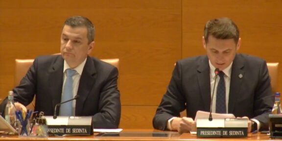 Two men in dark suits sit at a wood-paneled conference table with microphones and nameplates during a formal meeting. Left man looks ahead while right man reads notes into a microphone.