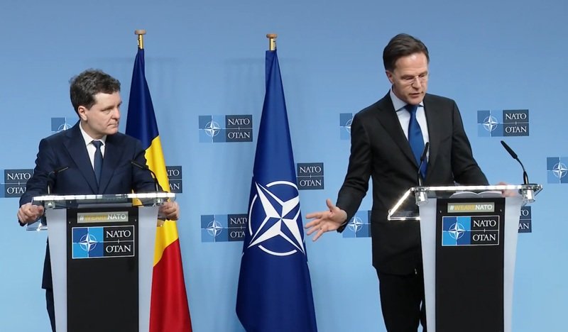 Two suited men stand at podiums during a NATO press conference, with a Romanian flag and NATO flag between them behind a blue backdrop marked NATO OTAN.