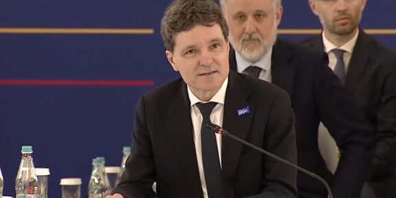 Man in a dark suit and tie speaking at a conference podium labeled 'ROMANIA' with a microphone and seated officials in the background.