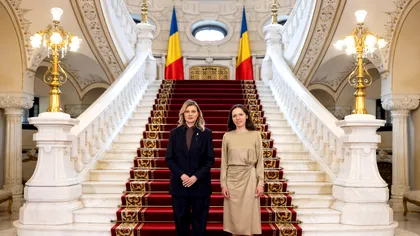 Two women in formal attire stand on a marble staircase in a government building, with Romanian flags in the background at the top of the stairs.