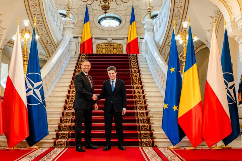 Two men in suits shake hands at a grand staircase, flanked by flags including Poland, NATO, EU, and Romania