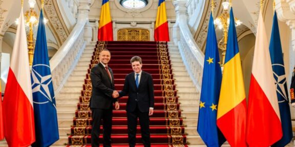 Two men in suits shake hands at a grand staircase, flanked by flags including Poland, NATO, EU, and Romania