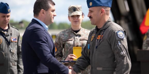 Civilian man in a dark suit shakes hands with a uniformed military officer during an award ceremony, with teammates in uniform in the background.