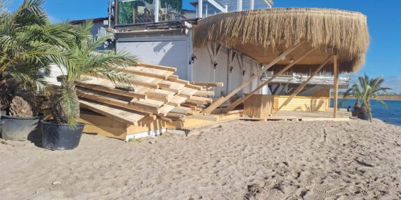 Stacks of wooden planks on a sandy beach beside a thatched-roof shed, with palm plants and a blue ocean in the background.