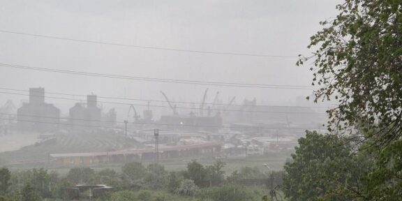 Industrial port and refinery structures with cranes visible through a hazy atmosphere, seen behind a curved railway track and greenery in the foreground.