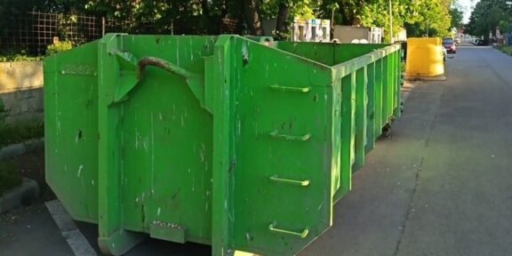Green dumpsters lined up along a city street, curbside with trees in the background and a yellow barrier in the distance.