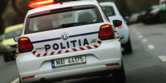 Rear view of a white Romanian police car with 'POLITIA' on the trunk and red emergency lights flashing on the roof, driving on a street.