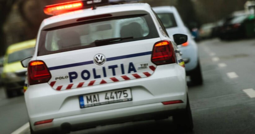 Rear view of a white Romanian police car with 'POLITIA' on the trunk and red emergency lights flashing on the roof, driving on a street.