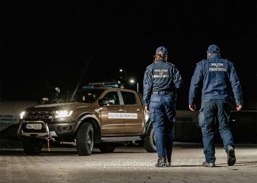 Two border police officers in uniform walk away from a patrol pickup at night, a 'Politia de Frontiera' vehicle nearby.