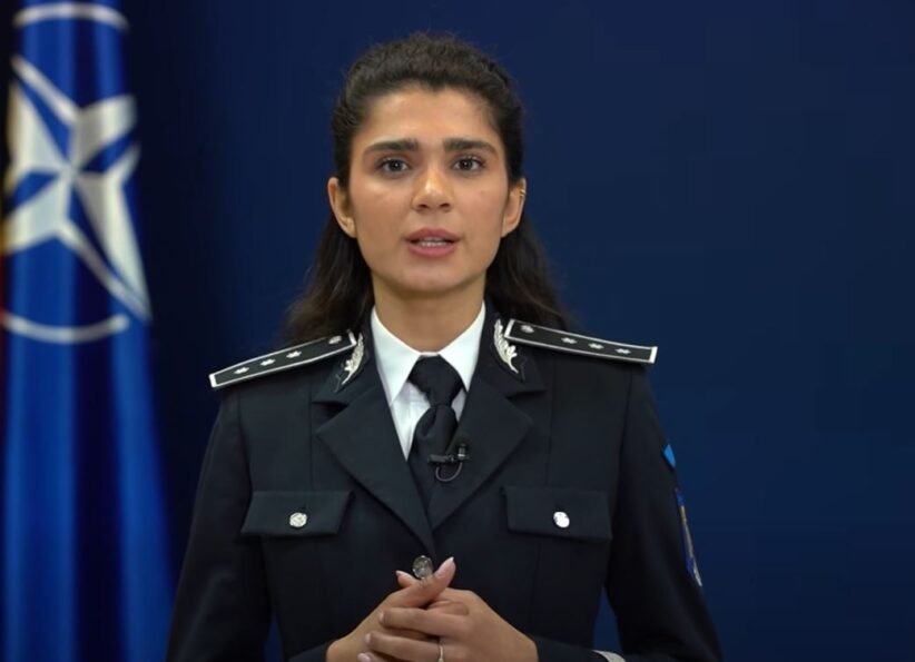Female police officer in uniform delivering a speech, hands clasped, with a blue NATO-style flag in the background.