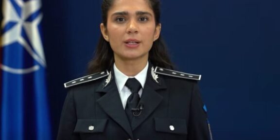 Female police officer in uniform delivering a speech, hands clasped, with a blue NATO-style flag in the background.
