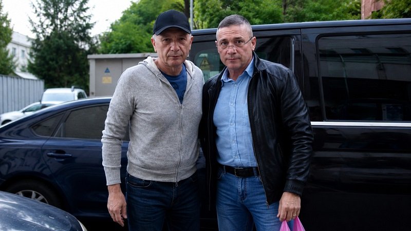 Two men pose together in front of a black van in a parking lot, dressed casually in jeans and jackets.