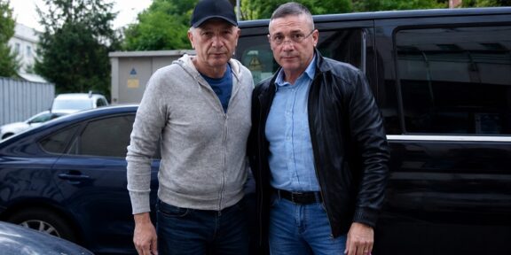 Two men pose together in front of a black van in a parking lot, dressed casually in jeans and jackets.