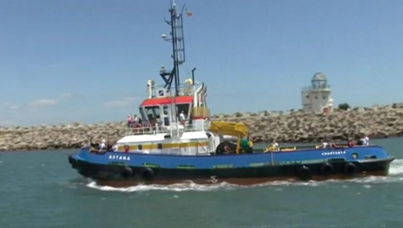 Blue tugboat Astana with a red-roofed cabin and yellow crane, sailing near a rocky breakwater on a sunny day in calm water.