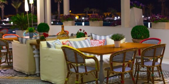 Nighttime outdoor cafe with wooden table and wicker chairs, cream couches, and potted plants on patterned tiled floor.