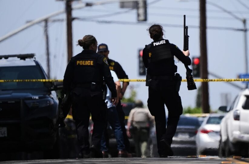 Police officers at a crime scene behind yellow tape, one officer holds a long firearm while another stands near a parked SUV.