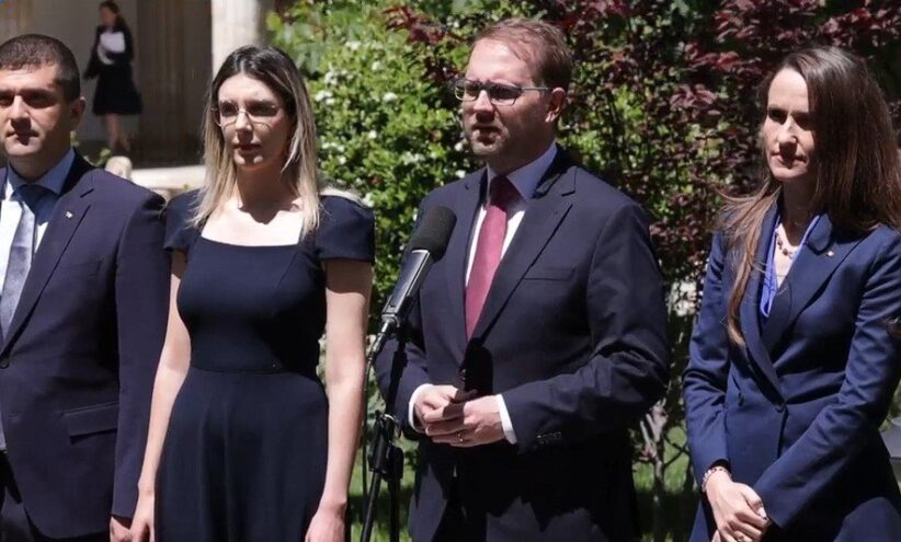 Four professionals in business attire stand outdoors; a man speaks into a microphone while three others listen nearby.