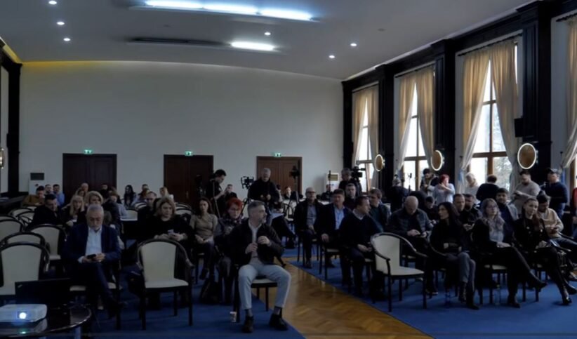 Audience seated in a large conference hall, awaiting a presentation; natural light from tall windows on the right side.