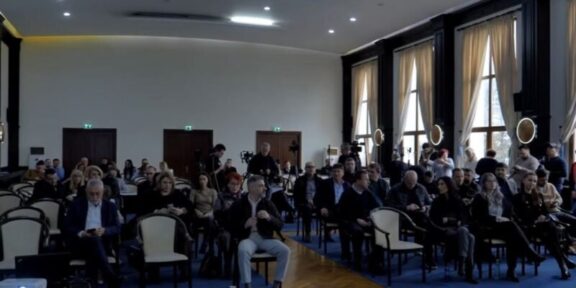 Audience seated in a large conference hall, awaiting a presentation; natural light from tall windows on the right side.