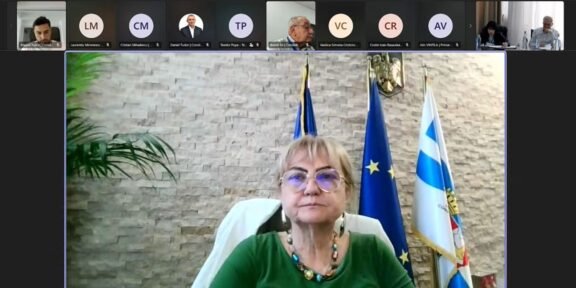 Framed woman in a green blouse wearing glasses, seated in a conference setting with European and flag banners behind her.