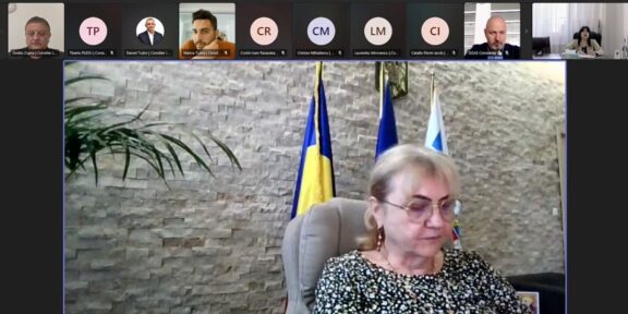 Video conference screenshot: a woman in a patterned blouse sits at a desk, with flags behind her and participants along the top thumbnails.