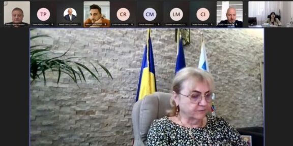 Video conference screenshot: a woman in a patterned blouse sits at a desk, with flags behind her and participants along the top thumbnails.